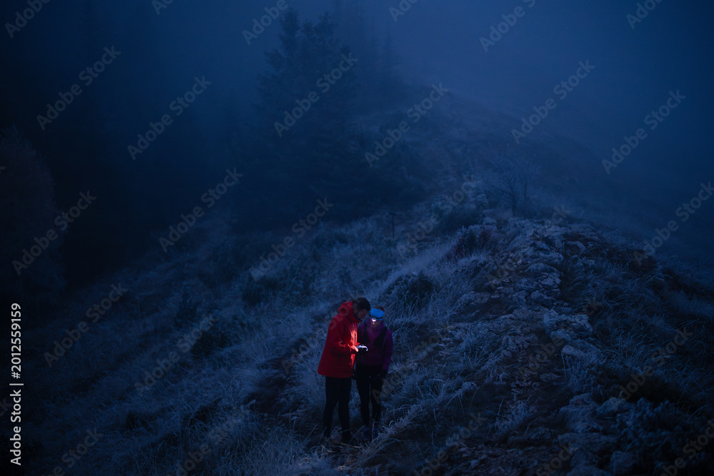 Naklejka premium Couple Hiking at Night on a Foggy Mountain Ridge Using Phone and Headlamp for Navigation