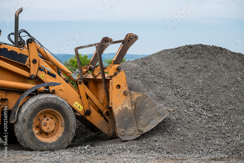 the front bucket of a yellow heavy machinery vehicle, such as a backhoe or wheel loader, pushing into a large mound of gray gravel.