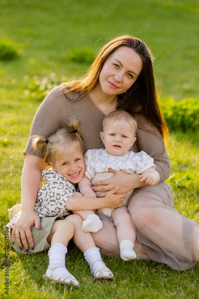 Fototapeta premium mother with two daughters sitting on a lawn at sunset in the summer, hugging