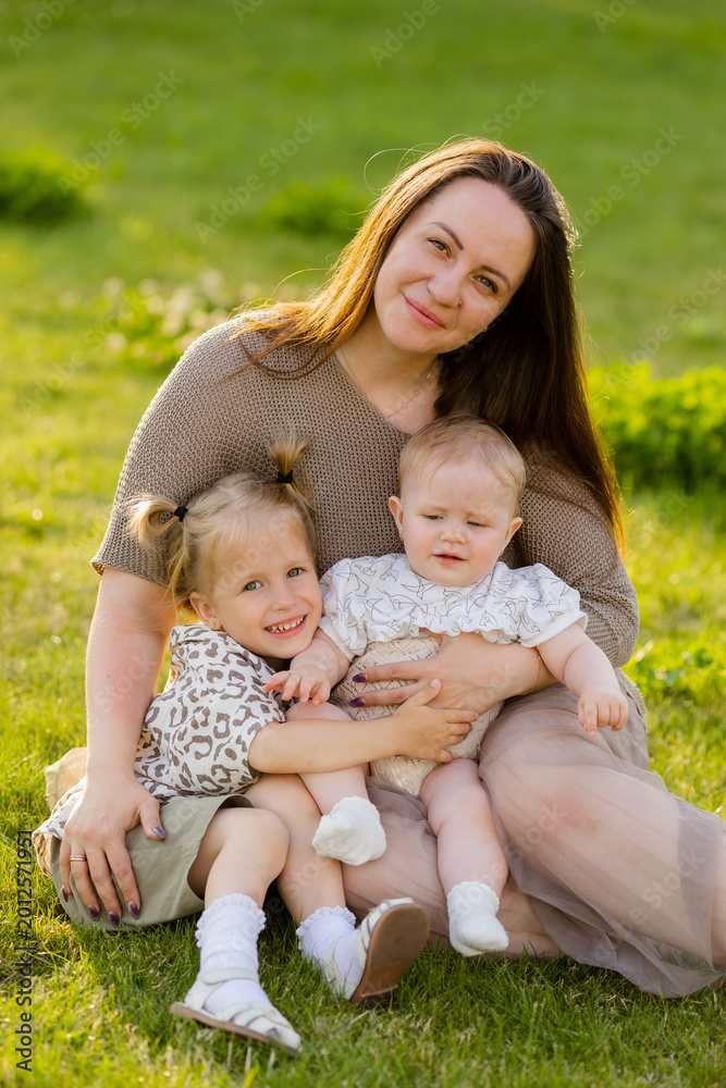 Fototapeta premium mother with two daughters sitting on a lawn at sunset in the summer, hugging