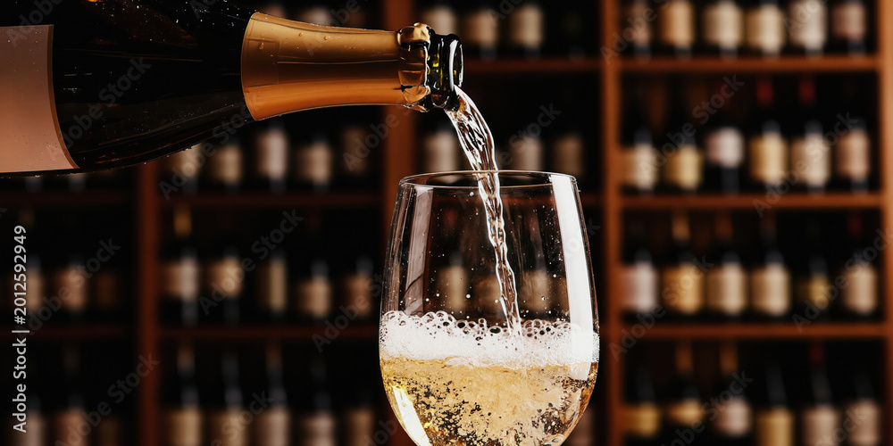 Fototapeta premium Sparkling wine poured from bottle into glass. Background shows cellar with many wine bottles. Bubbles form in glass creating festive mood. Perfect for celebrations and special events.