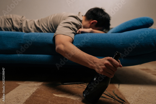 Photos Drunk man suffering from alcohol dependence, lying on blue sofa, holding bottle of alcoholic drink, depicting unconsciousness and devastating impact of substance misuse and long-term abuse