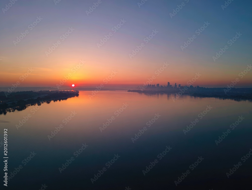 Obraz premium Sunrise over a tranquil river with a distant city skyline emerging from mist