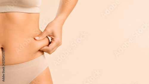 Close-up of woman's hand pinching waist fat, minimalist body contouring and slimming concept on beige background with massive copy space.