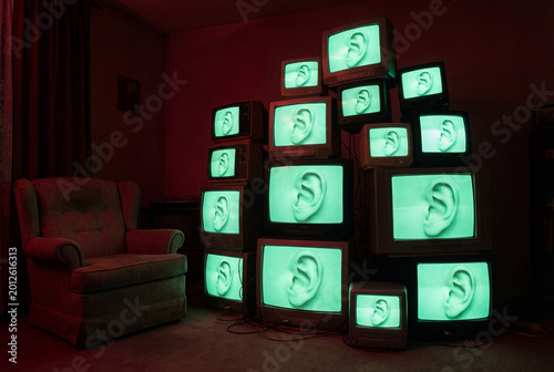Pile of vintage televisions in a dark red room next to an armchair. The glowing green screens all display a surreal close-up image of a human ear.