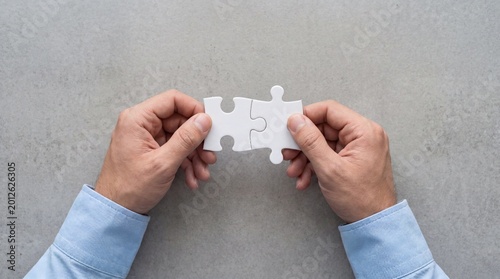 Photograph of hands holding two white jigsaw puzzle pieces about to connect, against a gray background.