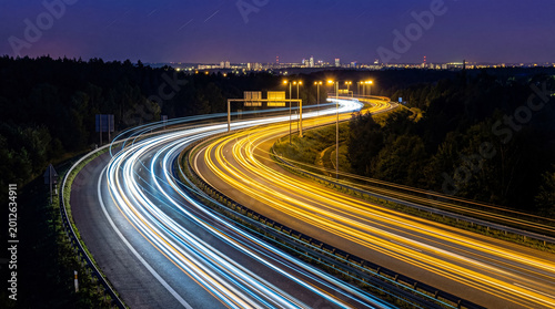 Fotografía A long exposure photograph of car light trails on a winding highway at night, with distant city lights and streetlamps