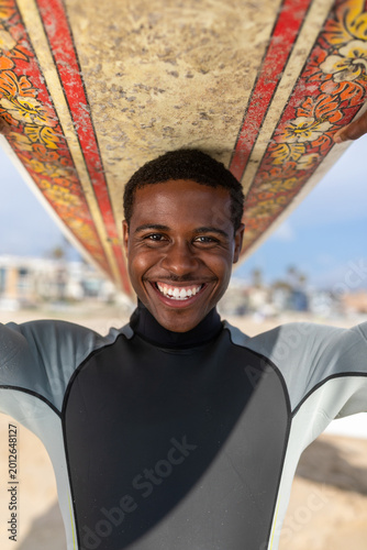 Canvastavla Smiling person in a wetsuit holding a surfboard overhead on a sunny beach, with buildings and blue sky in the background