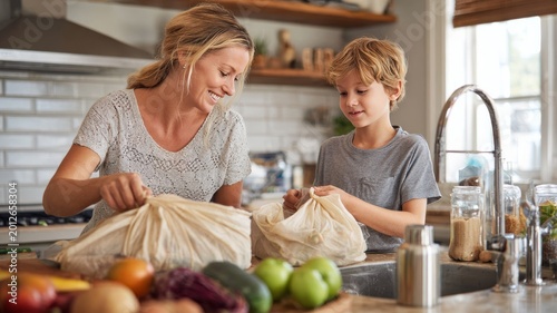 Mother and son unpacking reusable grocery bags filled with fresh produce in a bright kitchen
