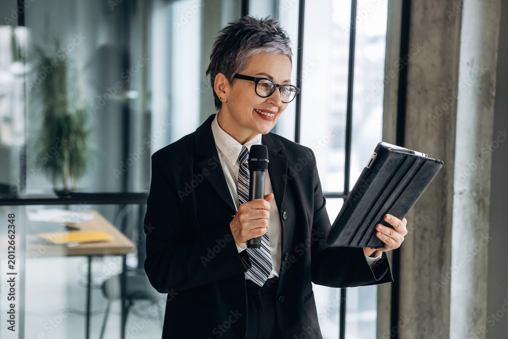 Fototapeta premium Businesswoman in formal attire holding a tablet and microphone while smiling, standing in a modern office with large windows and a contemporary workspace visible