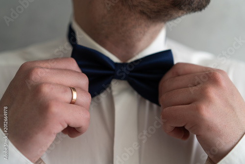 Close-up of a man adjusting a navy blue bow tie while wearing a gold wedding ring. A dapper groom or gentleman preparing for a formal ceremony, wedding, or elegant evening event.