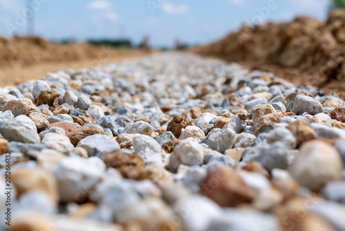 Fresh gravel path stretches toward distant horizon. Blurred dirt mounds frame the foreground. Clear sky with scattered clouds looms above. Construction site under development
