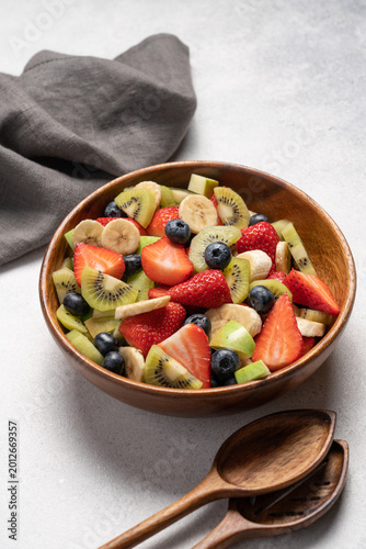 Fresh fruit salad with banana apple kiwi blueberry and strawberry in wooden bowl, top view on light background, healthy eating concept