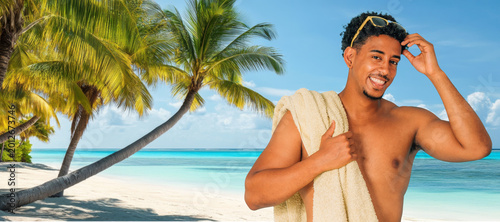 Young man smiling at a tropical beach, holding a towel over his shoulder, sunglasses on his head, with palm trees and blue ocean water