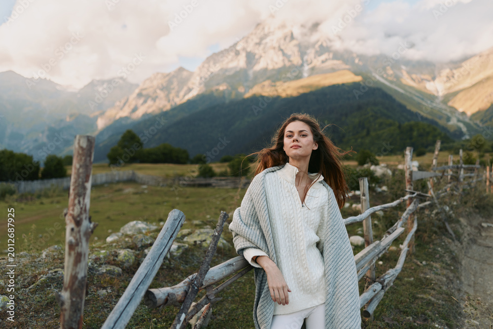 Naklejka premium A woman in a cardigan stands beside a wooden fence in a mountainside landscape, conveying fashion driven lifestyle and relaxed portrait in outdoor setting.