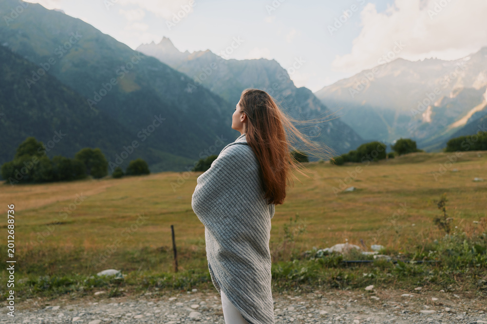 Naklejka premium Young woman wrapped in a gray shawl stands in a windswept meadow, looking toward distant mountains, conveying calm, wonder, and quiet connection with nature