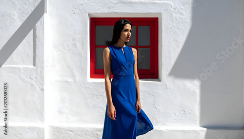 High-fashion women’s commercial, full-body shot from a three-quarter side perspective, outside a white stucco wall with a single red window, in bright natural sunlight casting soft, defined shadows.