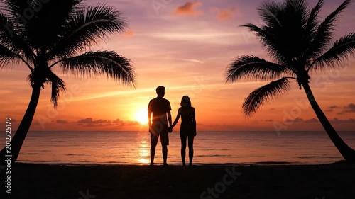Romantic couple holding hands and enjoying a stunning vibrant sunset on a tropical beach with a silhouetted palm tree, symbolizing love and peaceful vacation.