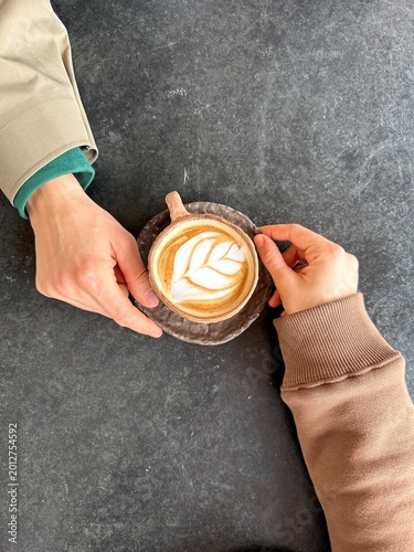 Top view of two hands holding a cup of coffee with latte art on a dark textured table