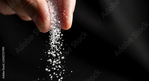 Close-up of fingers sprinkling coarse white salt. Salt crystals falling against a black background. Macro food seasoning concept