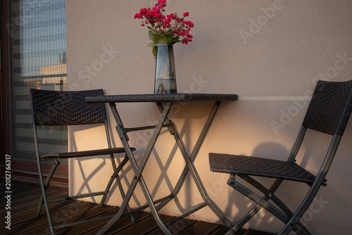 Cozy balcony terrace with bistro set and flowers in vase at sunset