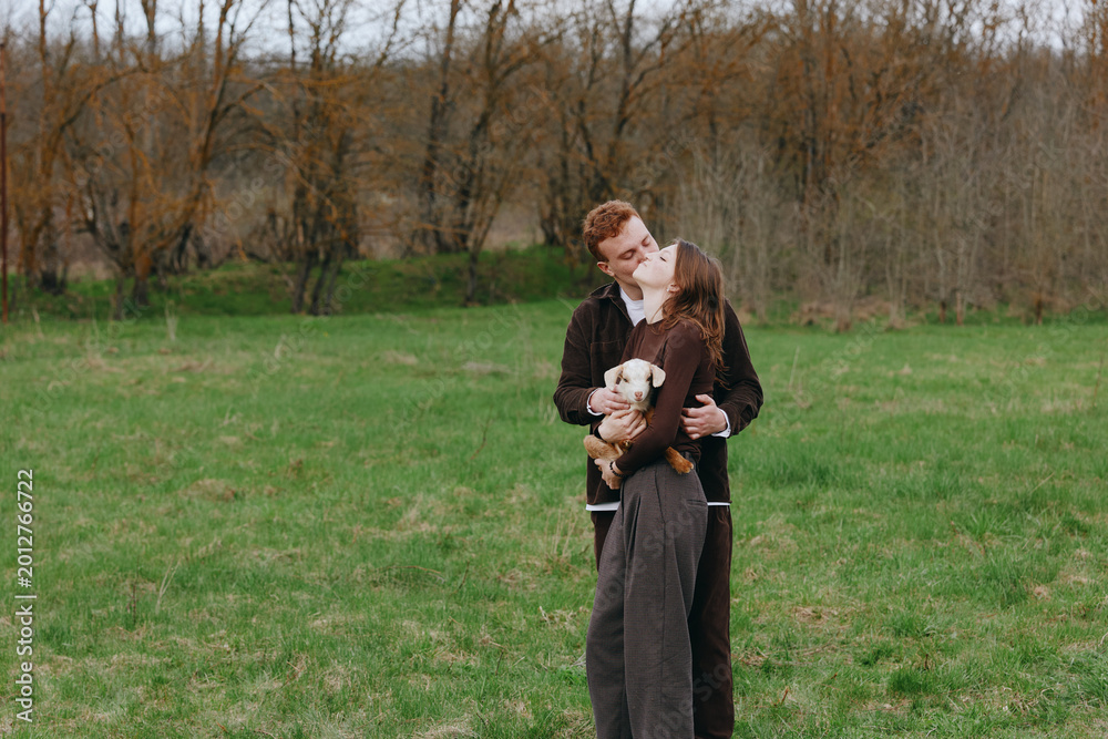 Obraz premium Young couple embraces tenderly in a green field while holding a small white goat, sharing a romantic moment in casual autumn clothing with trees in the background.