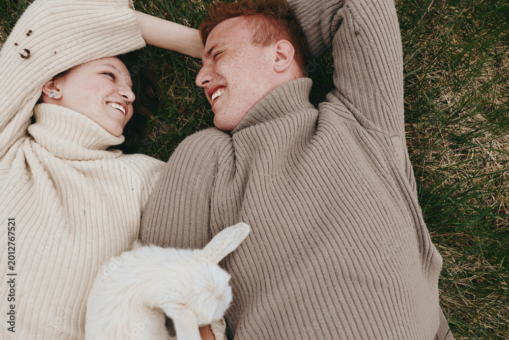 Naklejka premium Joyful couple outdoors, lying on grass with a playful goat kid nearby, cozy sweaters, sunlit scene, relaxed mood, candid affection, natural colors, serene countryside vibe