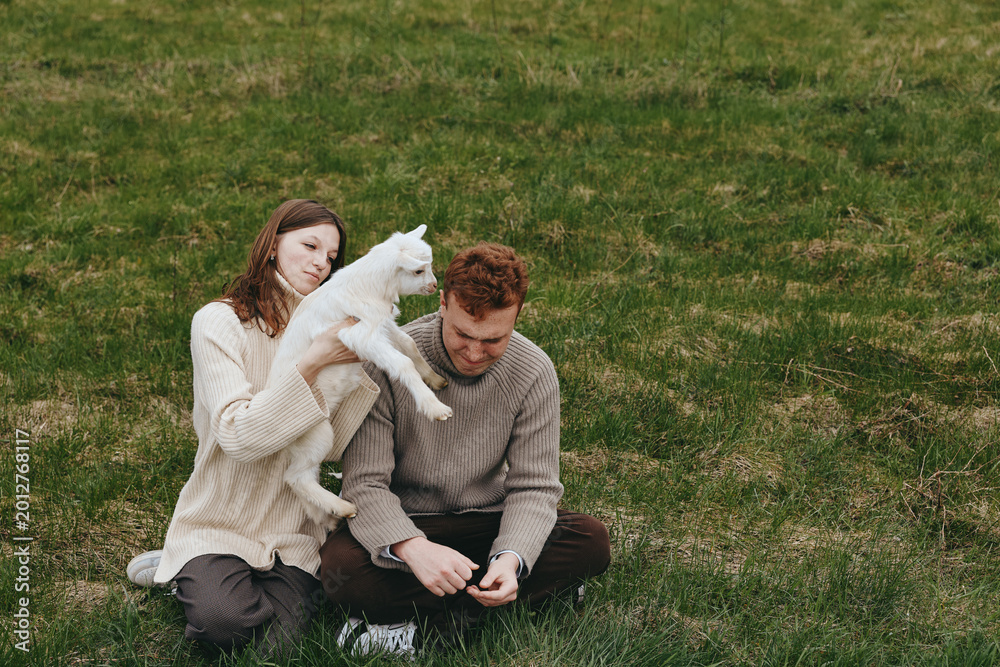 Naklejka premium Couple outdoors with a playful goat kid on a grassy field, capturing a warm moment of affection and connection. Natural light and relaxed mood emphasize lifestyle, care, and rural charm in this