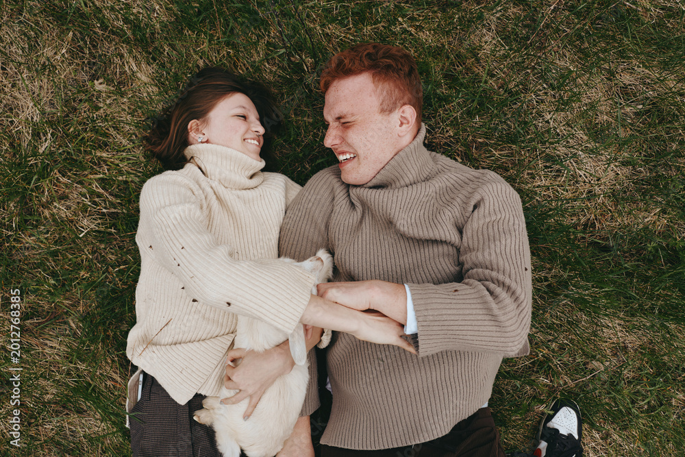 Naklejka premium Cheerful couple outdoors, lying on grass with a baby goat between them, sharing warm smiles, cozy sweaters, natural light, relaxed lifestyle moment in a lush green setting, full of affection