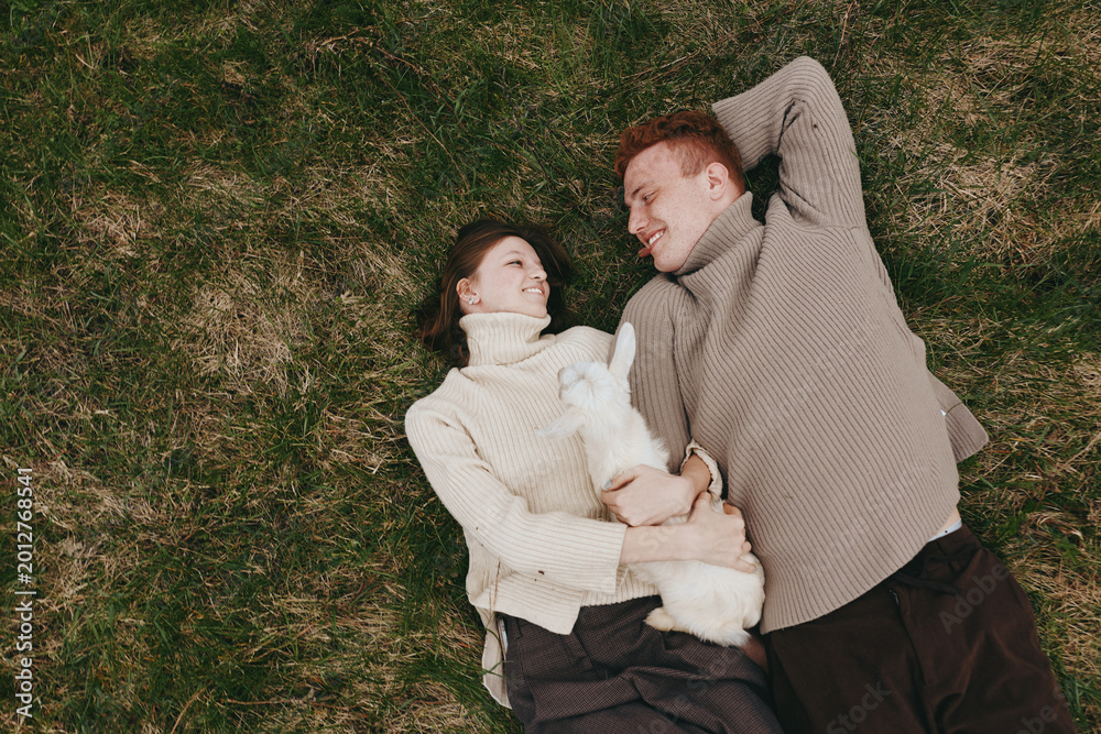 Obraz premium Couple lying on grass with a small white goat kid, cozy sweaters, and a gentle moment outdoors. Lifestyle scene highlighting bonding, family, and nature, with soft sunlight and relaxed, intimate mood.