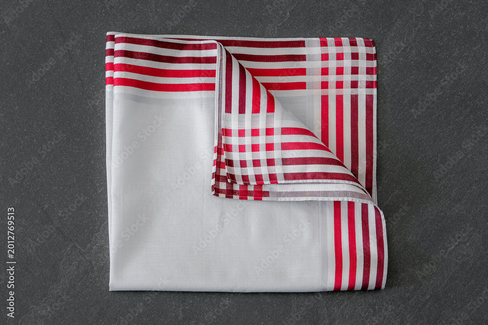Fototapeta premium A white cotton handkerchief with a bold red striped border and a checkered corner detail, neatly folded on a textured dark background.