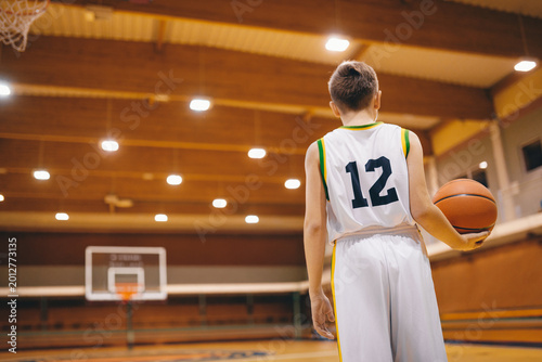 Junior Basketball Player in Number 12 Jersey Holding Ball on Indoor Court - Youth Athlete Standing in Gymnasium Prepared for Practice or Game