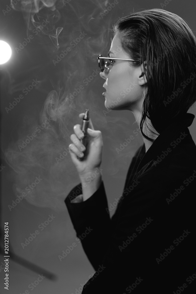 Fototapeta premium Black and white profile of a woman smoking, wearing glasses and dark clothing in a moody atmosphere.