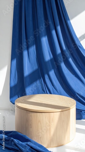A round wooden table set against a backdrop of flowing blue fabric, illuminated by natural sunlight in a contemporary space.