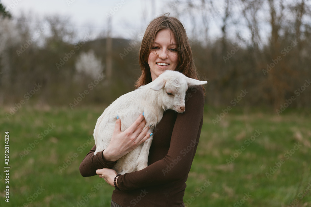 Naklejka premium A smiling woman outdoors holds a young goat kid in a grassy field, presenting a warm, gentle lifestyle moment. The scene emphasizes bonding, care, and natural companionship between human and animal.
