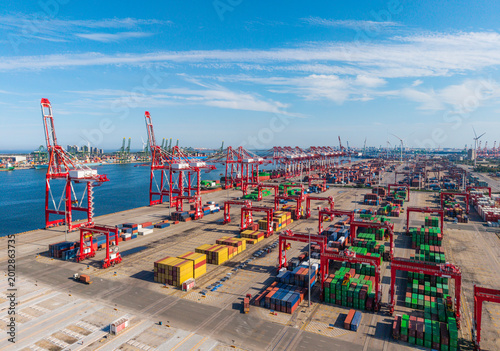 Large port container terminal with cranes and stacked shipping containers under clear blue sky