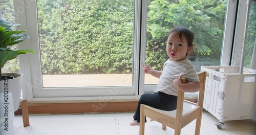 Free Download A curious Asian toddler sits on a wooden chair in a sunlit room, observing her environment in a moment of quiet learning and early childhood development. - GFXLoad