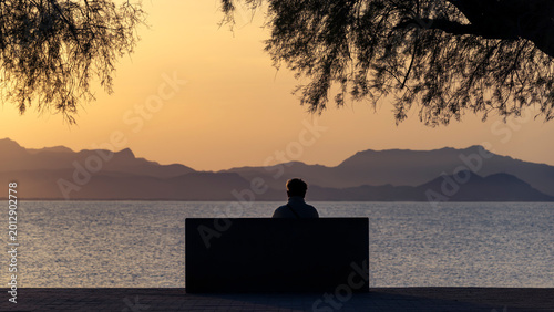 Mallorca seascape and landscape, with silhouetted and unrecognizable person sitting on a bench, looking at the mediterranean sea and Serra de Tramuntana in the distance.