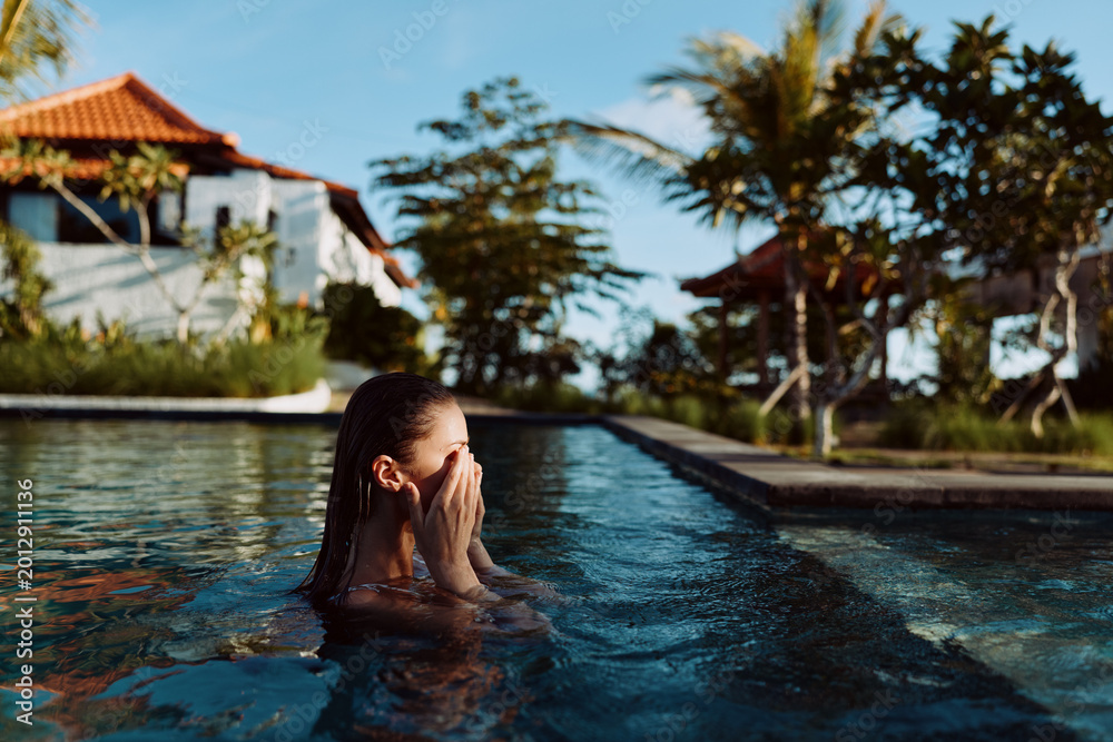 Fototapeta premium Young woman with wet hair covering her face while standing in an outdoor pool. Tropical trees and houses in the background. Concept of relaxation and lifestyle.