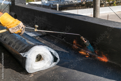 Fotografía a professional roofer applying heat to a waterproof bitumen membrane on a flat commercial rooftop using a gas blowtorch