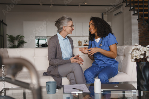 Konstfotografi Elderly woman and nurse having conversation while sitting on couch at home, medi