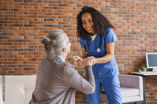 Female caregiver helping elderly woman stand up at home , young african-american nurse holding senior patient by hand, home care, rehabilitation support and mobility assistance concept