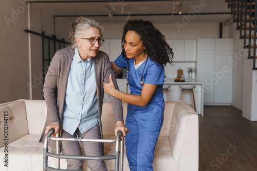 Senior woman walking with walker while nurse helps her at home, caregiver supporting elderly patient with mobility assistance, rehabilitation, home care service and healthcare concept copy space