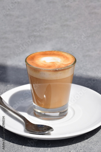 A small glass cup of cappuccino on a white plate with a silver spoon.