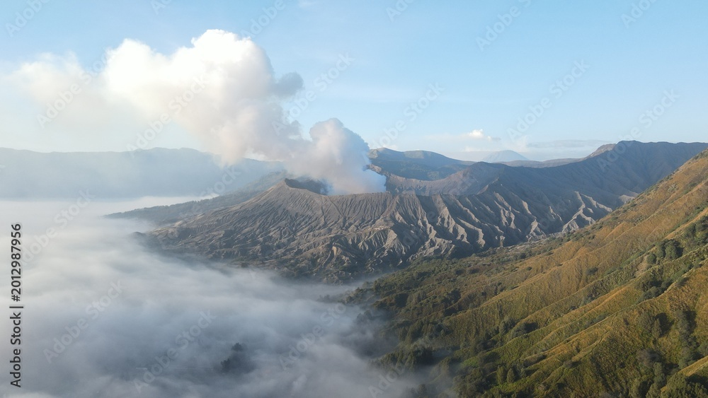 Obraz premium Stunning aerial view of Mount Bromo volcano erupting with white smoke above a thick morning mist.