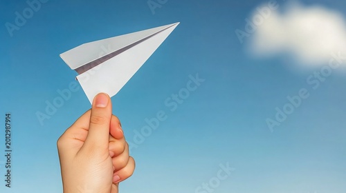 Human hand holding small simple white paper airplane ready to launch against clear soft blue sky with single fluffy cloud in gentle background