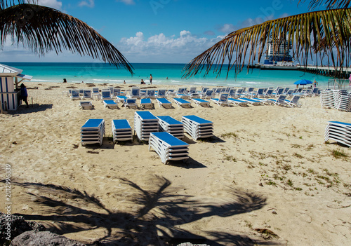 Blue beach lounge chairs stacked at a tropical beach. There are tourists and a cruise ship in the background