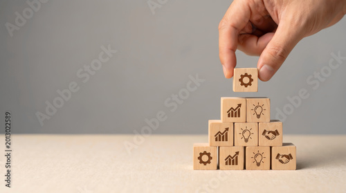 Business strategy and growth management planning concept with hand stacking wooden blocks featuring icons for success innovation partnership and leadership development on grey background.