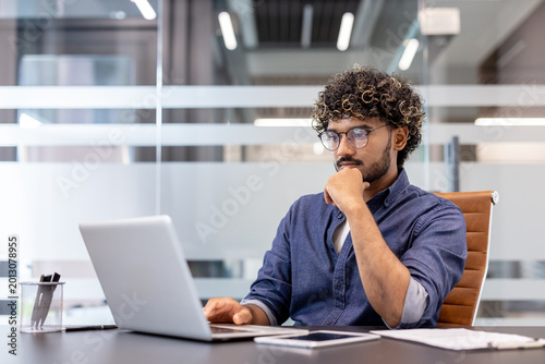 A thoughtful young Indian man is sitting at a desk in the office and looking at the laptop screen
