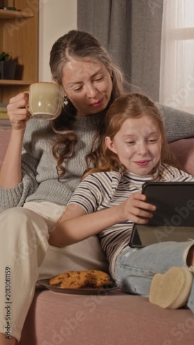 Vertical shot of adult woman drinking coffee while watching film on tablet with preteen daughter, both lying back on couch in living room